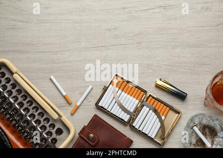 Offenes Gehäuse mit Tabakfilterzigaretten, Feuerzeug, Aschenbecher aus Glas und Brieftasche in der Nähe einer alten Schreibmaschine auf einem Holztisch, flach liegend. Platz für Text Stockfoto