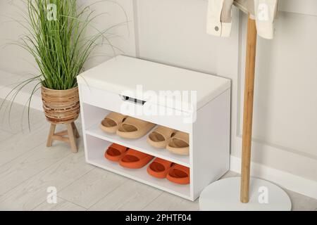 Elegante Aufbewahrungsbank mit verschiedenen Paar Gummipantoffeln und wunderschönen Zimmerpflanzen in der Nähe der weißen Wand im Flur Stockfoto