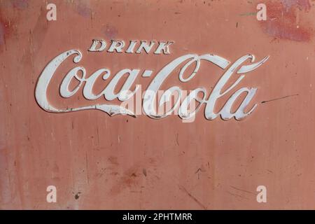 Verschluss des Kühlschranks mit den Worten: COCA-COLA IN weißen Buchstaben TRINKEN. Verblasste rote Seite mit Streifen, Rost und einem sehr verwitterten Vintage-Look. Stockfoto