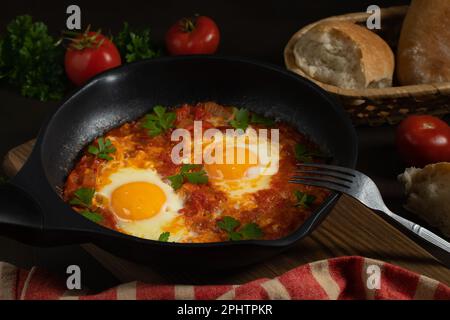 Shakshuka aus zwei Eiern in Tomatensoße mit frischen Tomaten, Gewürzen und Kräutern in einer schwarzen Pfanne. Rührei aus der Nähe. Stockfoto