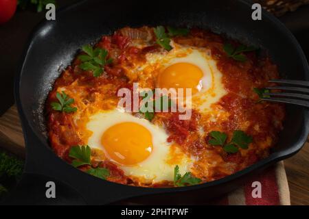 Shakshuka aus zwei Eiern in Tomatensoße mit frischen Tomaten, Gewürzen und Kräutern in einer schwarzen Pfanne. Rührei aus der Nähe. Stockfoto