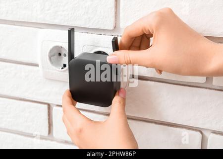 Frau, die den schwarzen WLAN-Repeater in die Steckdose an der weißen Backsteinwand steckt Stockfoto