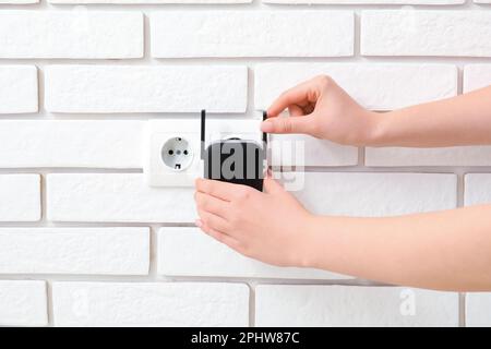 Frau, die den schwarzen WLAN-Repeater in die Steckdose an der weißen Backsteinwand steckt Stockfoto