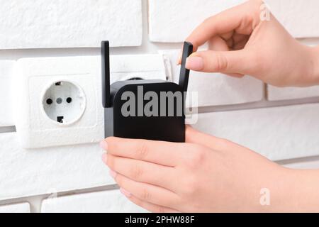 Frau, die den schwarzen WLAN-Repeater in die Steckdose an der weißen Backsteinwand steckt Stockfoto