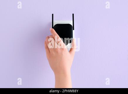 Frau, die den schwarzen WLAN-Repeater in die Steckdose an der lilafarbenen Wand steckt Stockfoto