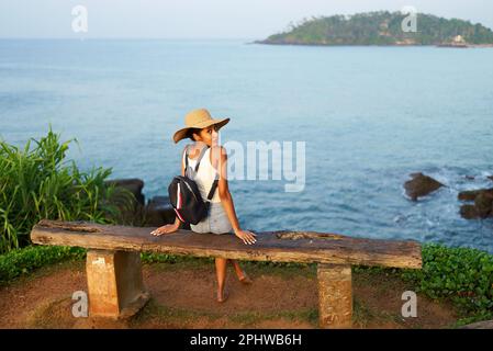 Eine multiethnische Frau mit Strohhut genießt einen tropischen Urlaub auf einer Bank an der Klippe mit Blick auf das Meer. Schwarze Frau mit Rucksack für Besichtigungstouren auf landschaftlich reizvoller Landschaft Stockfoto