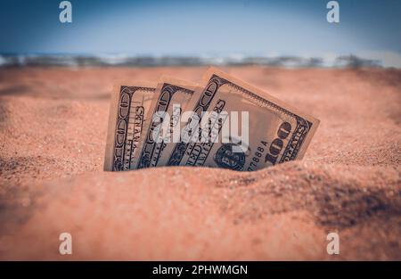 Die mit Sand bedeckten Gelddolare liegen am Sandstrand in der Nähe der Meereswellen am sonnigen Sommertag aus nächster Nähe. Geld wächst aus dem Boden. Konzept Finanzierung Geld Urlaub Relax Urlaub. Stockfoto