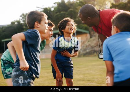 Teammotivation auf einem Grundschulgelände. Sporttrainer, der während des Sportunterrichts mit einer Gruppe von Grundschulkindern aufmunternd spricht. Sport und Fitness t Stockfoto