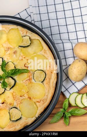 Eine Quiche mit Basilikumblättern, Kartoffeln und Zucchini in Scheiben auf einem Holzbrett mit ganzen Kartoffeln daneben Stockfoto