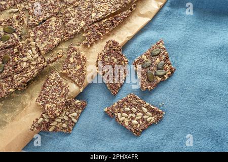 Knusprige Vollkorn-Snacks aus Hafer und Leinsamen garniert mit verschiedenen Samen auf Backpapier und einer blauen Serviette, gesundes Backen, Draufsicht von oben, Kopie Stockfoto