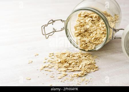 Haferflocken fallen aus einem Glasgefäß auf einen hellen Holztisch, gesunde Ernährung, Kopierraum, ausgewählter Fokus, schmale Schärfentiefe Stockfoto