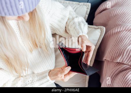 Unglückliche junge Frau, die zu Hause warme Kleidung trägt und eine leere Brieftasche in der Hand hat. Energiekrise in Europa infolge von Inflation und Krieg. Preiserhöhung für Stockfoto