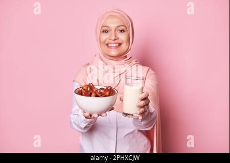 Eine Schüssel mit Datteln und einem Glas Milch, in den Händen einer lächelnden muslimischen Frau im Hijab, AUF pinkfarbenem Hintergrund. Ramadan Kareem Stockfoto