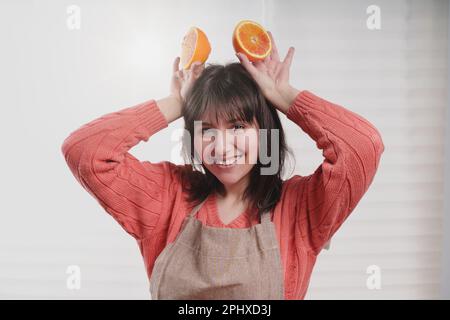 Fröhliche junge Weiße, trägt eine Schürze über ihrem Pulli, hält ihr orangefarbene Hälften an den Kopf wie Mäusenohren, lächelt und schaut auf den Camer Stockfoto