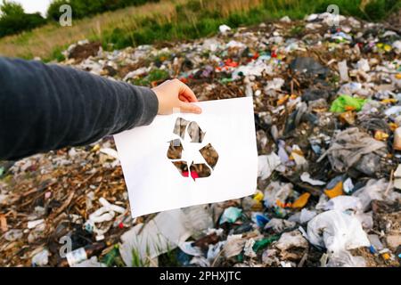 Müllverschmutzung. Handgehaltenes, geschnittenes Papier mit dem Logo „Recycling“ auf Müllkippen-Müll-Hintergrund. Recycling-Konzept. Verschmutzung des Planeten. Raus Stockfoto