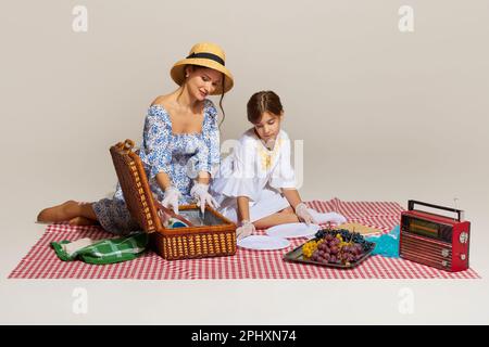 Foto einer wunderschönen Mutter mit einer kleinen Tochter, die das Essen aus dem Korb nimmt, vor hellem Hintergrund. Glückliche Familie beim Picknick Stockfoto