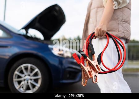 Nahaufnahme eines Autovoltmeters in den Händen einer Frau vor dem Hintergrund eines kaputten Autos Stockfoto