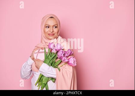 Eine wunderschöne Frau in pinkem Hijab, lächelnd zur Seite, mit einem Blumenstrauß mit lila Tulpen und einer Geschenkbox, isoliert auf rosa Stockfoto