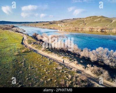Draufsicht auf einen Sportler, der bei Sonnenuntergang über eine staubige Landstraße in der Nähe des Flusses und der Hügel läuft. Wanderweg an der Steppe in der Nähe des Flusses Ili im Süden Stockfoto