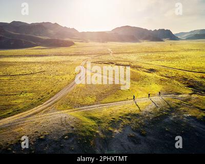 Draufsicht über drei Sportler, die auf einer staubigen Landstraße in grünen, grasbedeckten Hügeln mit felsigen Bergen bei Sonnenuntergang unterwegs sind. Traillauf A Stockfoto