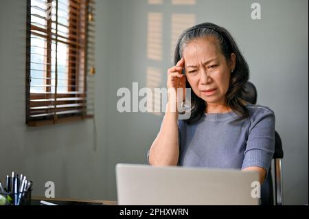 Verwirrte und unglückliche Frau im asiatischen Alter sitzt an ihrem Schreibtisch, hat ein Problem mit ihrer Internetverbindung auf ihrem Laptop oder hat Probleme mit ihrem B-B. Stockfoto