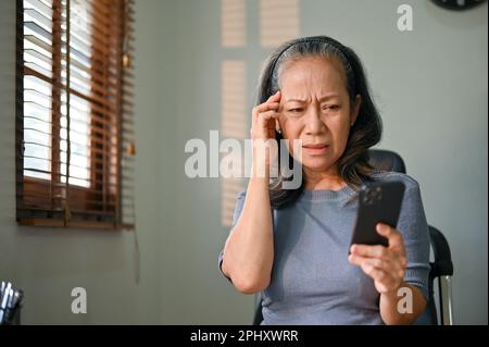 Verwirrte und unglückliche Frau im asiatischen Alter sitzt an ihrem Schreibtisch und hat ein Problem mit ihrer Internetverbindung auf ihrem Smartphone, und sie hat Kopfschmerzen mit Technol Stockfoto