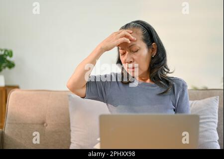 Gestresste ältere Asiatin berührt ihre Stirn, leidet an Kopfschmerzen oder Migräne, während sie zu Hause mit einem Notebook an ihren geschäftlichen Aufgaben arbeitet. Stockfoto