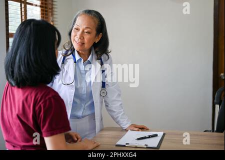 Professionelle Senior asiatische Ärztin konsultiert einen medizinischen Behandlungsplan mit einem Patienten im Untersuchungsraum einer Klinik. Stockfoto