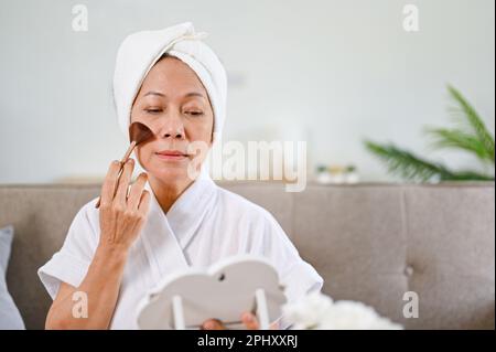 Eine schöne Frau im asiatischen Alter im Bademantel, die sich schminkt, ihre Wange mit einer Bürste besticht und in den Spiegel schaut. Stockfoto