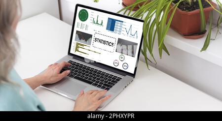 Die Analystin an ihrem Schreibtisch arbeitet an einem Laptop, der Statistiken, Diagramme und Diagramme zeigt. Sie arbeitet am Holztisch im Kreativbüro. Über die Stockfoto