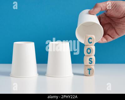 Das Konzept der versteckten Kosten finden. Ein Mann, der das Hütchenspiel mit drei Tassen spielt, enthüllt das Wort "Kosten" auf Holzwürfeln. Stockfoto
