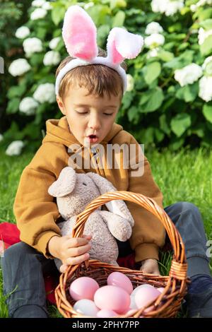 Der kleine weiße Junge trägt Kaninchenohr und sammelt ostereier in einem Korb auf einer ostereiersuche im Garten Stockfoto