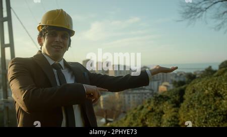Innovativer und professioneller junger Bauingenieur oder Auftragnehmer in Anzug und Helm, der moderne Wohnanlagen in städtischer Landschaft präsentiert Stockfoto