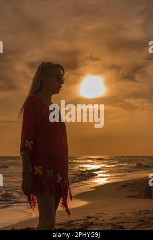 Seniorin steht am Strand während des Sonnenuntergangs. Sommer- und Urlaubskonzept. Stockfoto