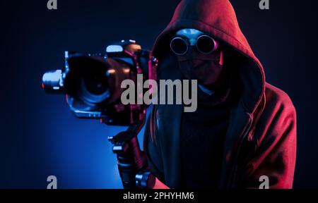 Ein Typ in Kapuze mit Kamera. Neonbeleuchtung. Der junge europäer ist im Dark Studio Stockfoto