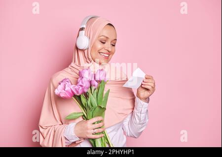 Entzückende arabische muslimische Frau in pinkfarbenem Hijab, liest Grußkarte, während sie einen Strauß Tulpen zum Muttertag bekommt Stockfoto