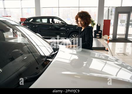 Lächelnde, lockige Kundin legte ihre Hand auf ein funkelndes Auto mit Haube Stockfoto