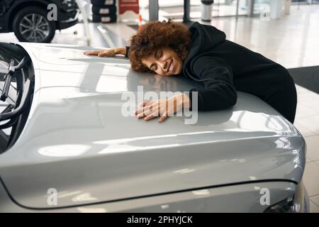 Glückliche multirassische Frau, die die Motorhaube eines neuen Autos umarmt Stockfoto