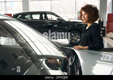 Eine lächelnde, multirassische Frau legte ihre Hand auf die Motorhaube eines neuen Autos Stockfoto