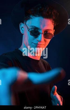 Mit Hut und Brille. Neonbeleuchtung. Der junge europäer ist im Dark Studio Stockfoto