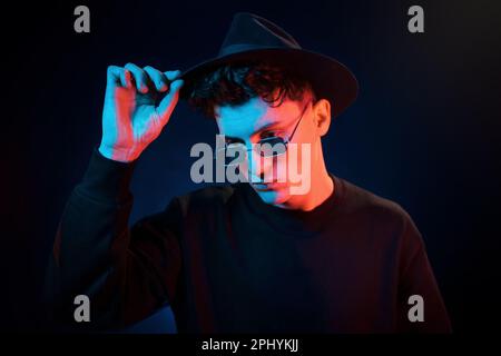 Mit Hut und Brille. Neonbeleuchtung. Der junge europäer ist im Dark Studio Stockfoto