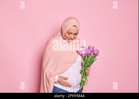 Entzückende muslimische schwangere Frau in rosa Hijab, berührt den Bauch, drückt positive Emotionen aus, spürt das erste Baby tritt Stockfoto