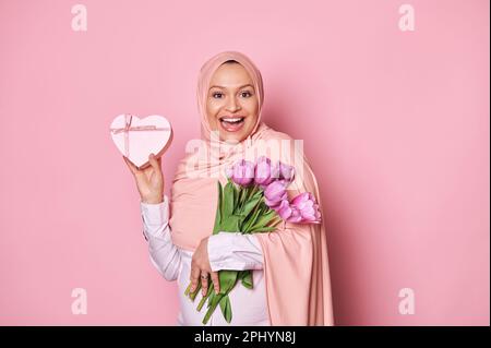 Muslimische schwangere Frau in pinkfarbenem Hijab, mit einem Strauß lilafarbener Tulpen und einer herzförmigen Geschenkbox zum Muttertag Stockfoto