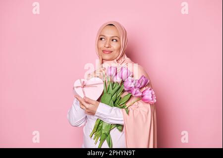 Pensive muslimische schwangere Frau im Hijab, träumlich zur Seite blickend, mit einem Strauß aus Tulpen und einer herzförmigen Geschenkbox in der Hand, Stockfoto