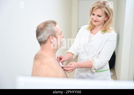 Lächelnde Ärztin mit holter-Herzmonitor auf der Brust des Patienten Stockfoto