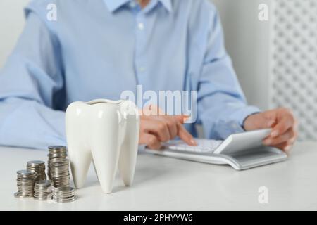 Frau mit Taschenrechner am weißen Tisch in Innenräumen, Fokus auf das Keramikmodell von Zahn und Münzen. Teure Behandlung Stockfoto