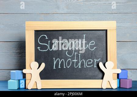 Kleine Tafel mit der Phrase „Surrogate Mother“, Figuren und Würfeln auf blauem Holztisch Stockfoto