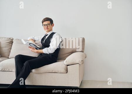 Ein junger Mann in formeller Kleidung sitzt auf dem Sofa drinnen an einer weißen Wand Stockfoto