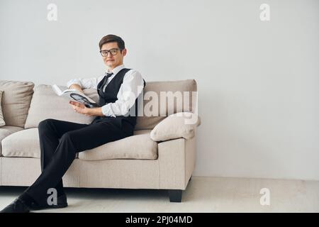 Ein junger Mann in formeller Kleidung sitzt auf dem Sofa drinnen an einer weißen Wand Stockfoto