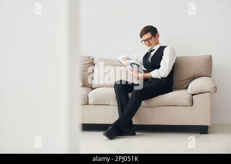 Ein junger Mann in formeller Kleidung sitzt auf dem Sofa drinnen an einer weißen Wand Stockfoto